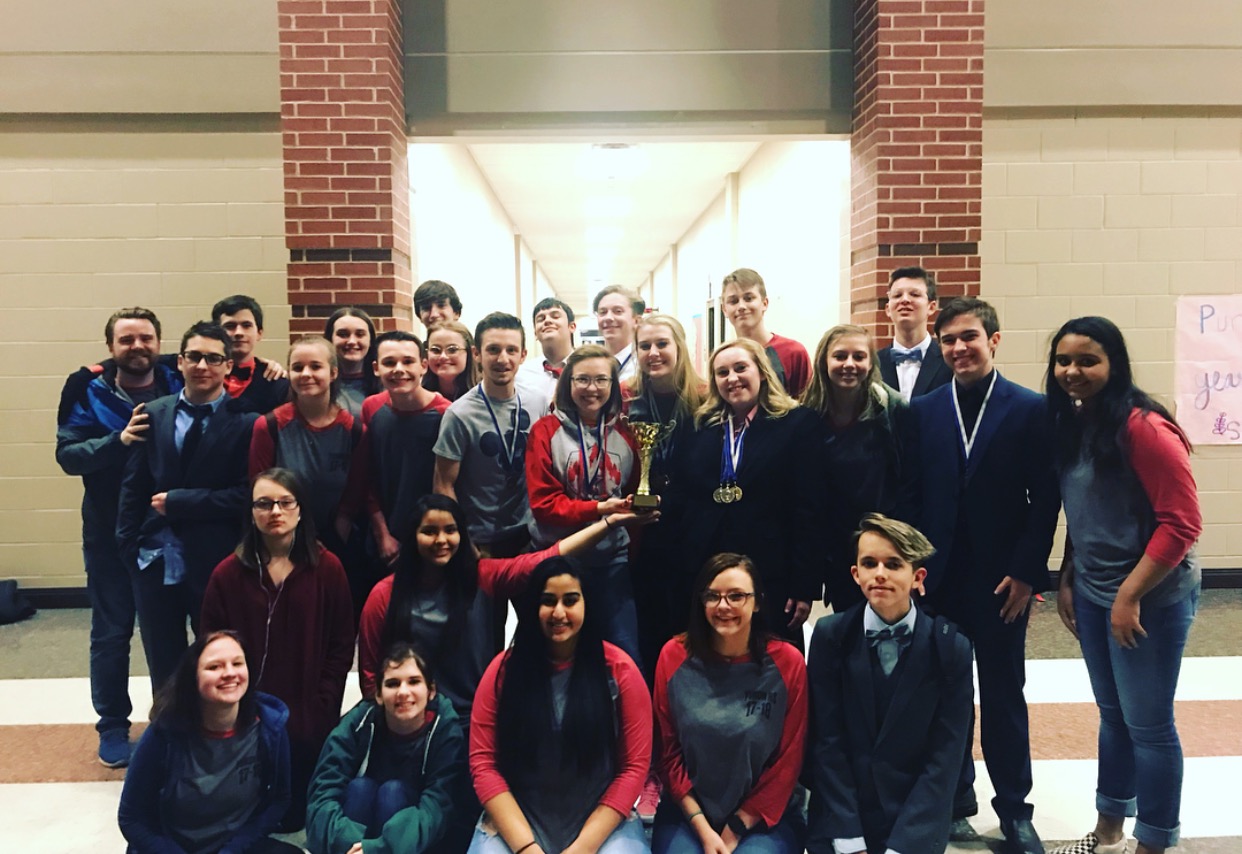 A large group of students poses together in a school hallway, some holding trophies and wearing medals, celebrating a recent achievement.