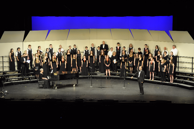 A large choir dressed in black performs on stage, with a conductor leading and a pianist seated at a piano, set against a blue backdrop.