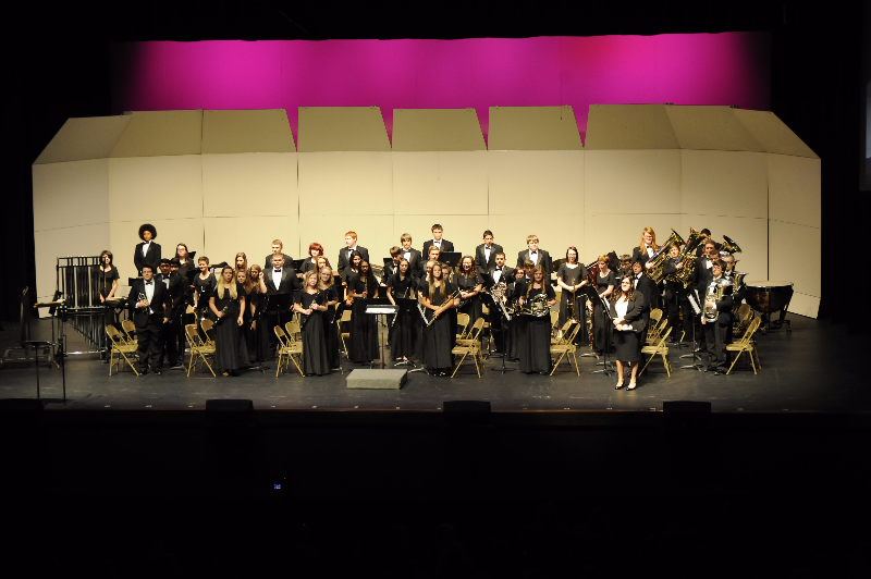 A large group of musicians dressed in formal attire stands on stage, arranged in rows with various instruments, against a backdrop of pink lighting and a simple set design.