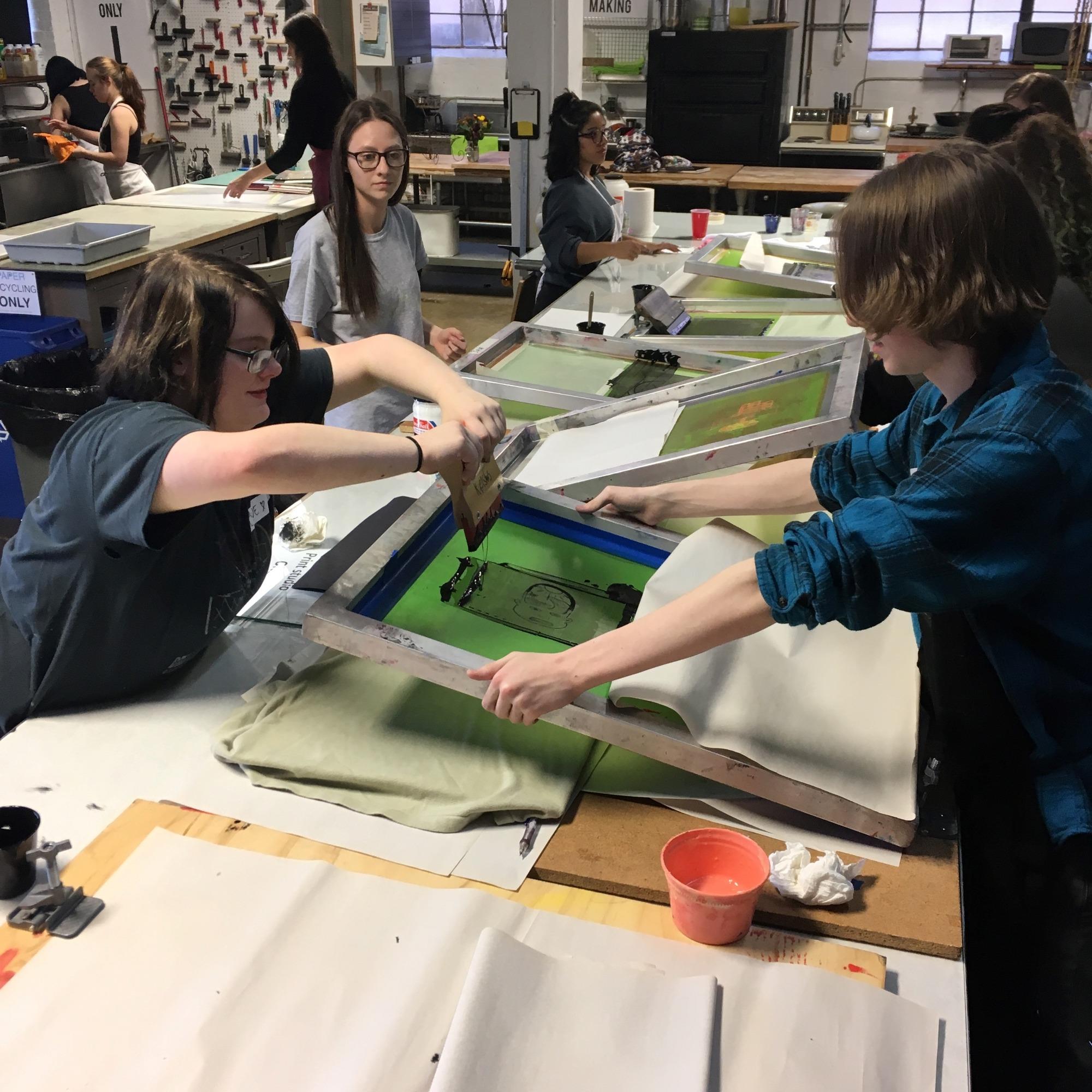 Two individuals are collaboratively screen printing in an art studio, surrounded by various materials and other people engaged in similar activities.