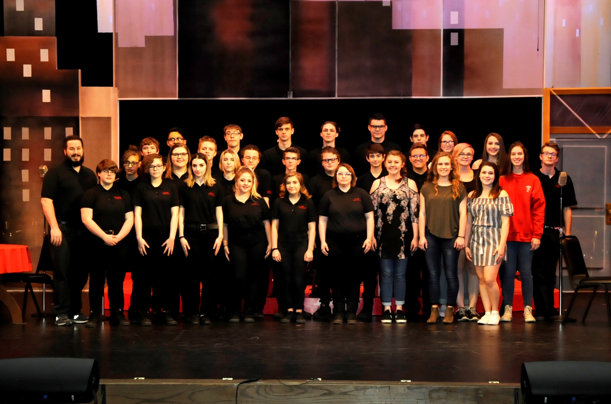 A group of students and instructors pose together on stage, dressed in black shirts, with a colorful backdrop of city buildings behind them.