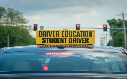 A rear view of a car displaying a bright yellow sign that reads 'DRIVER EDUCATION STUDENT DRIVER' on top, with traffic lights visible in the background.