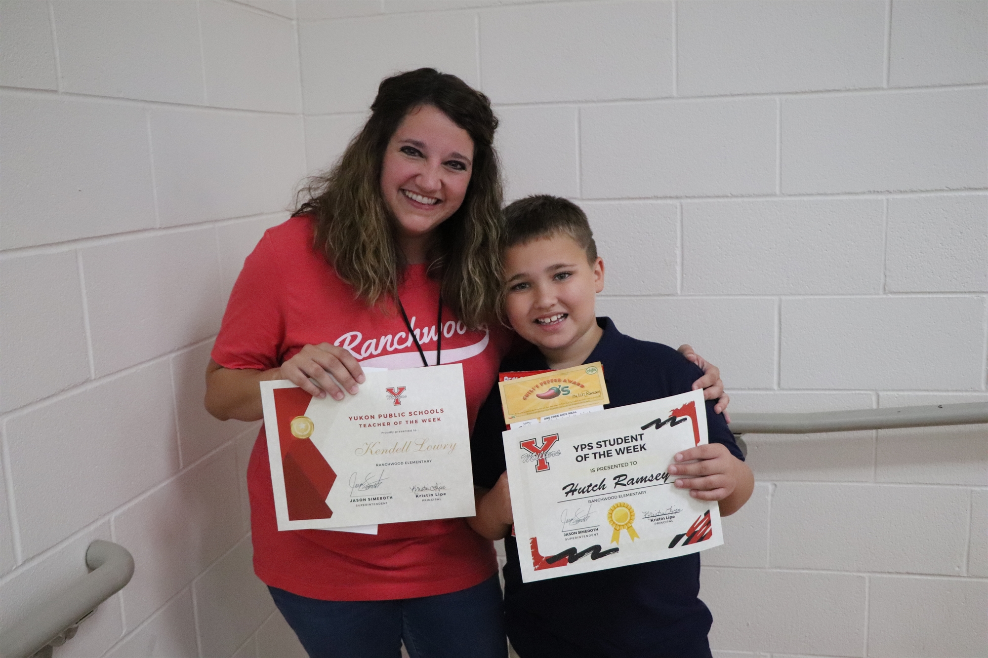 A woman and a boy stand together, both smiling and holding certificates, in a hallway with white walls.