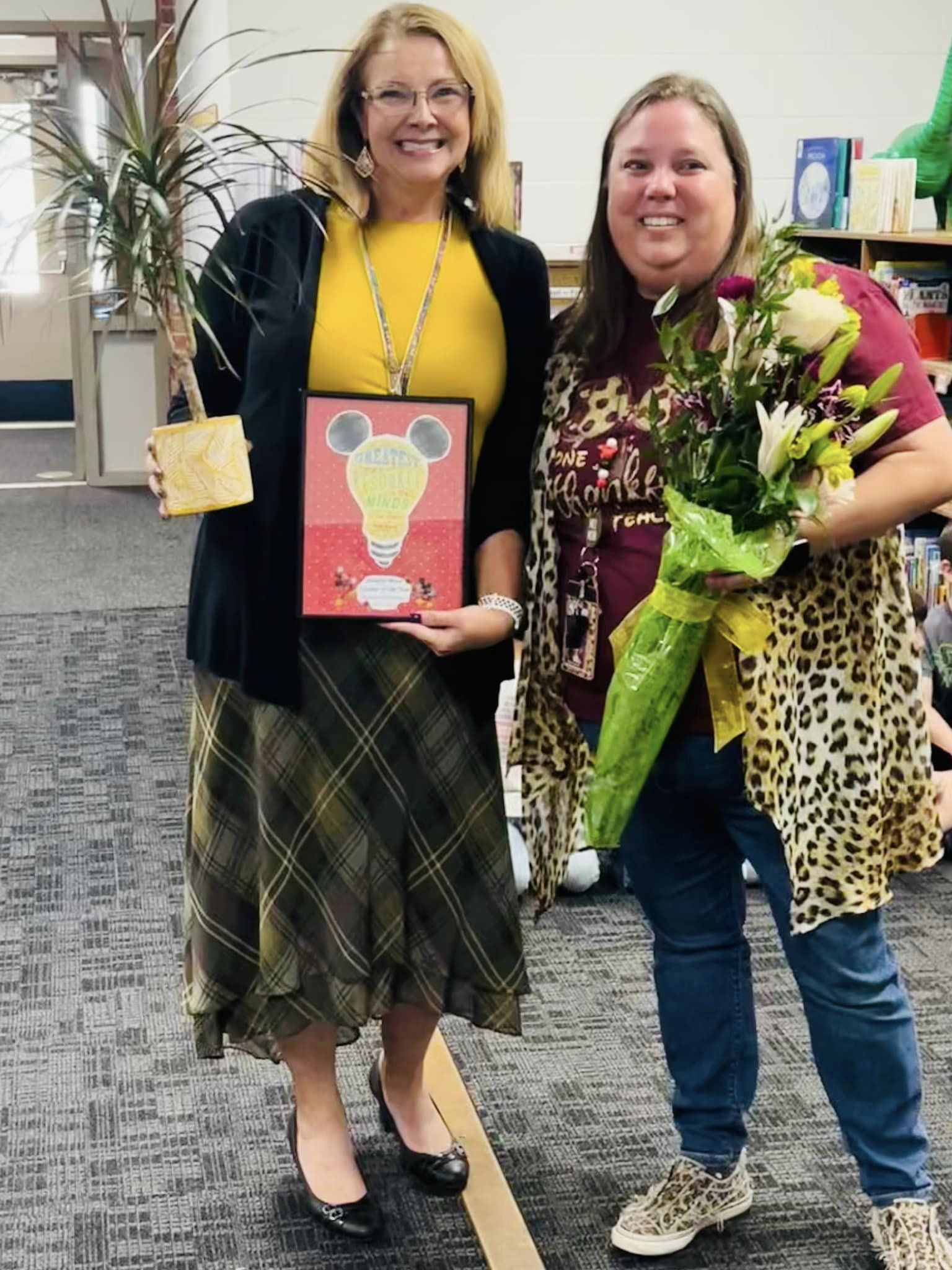 Two women stand together in a classroom setting, one holding a framed certificate and the other holding a bouquet of flowers, both smiling at the camera.
