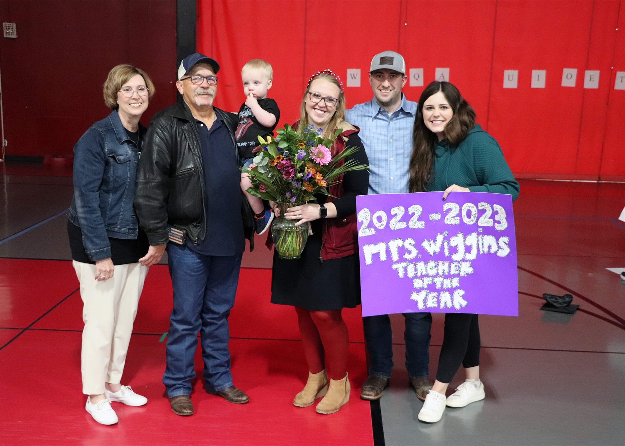 A group of five people, including a woman holding a bouquet of flowers and a sign that reads '2022-2023 MTS Winds Teacher of the Year,' celebrate in a gymnasium with a red backdrop.