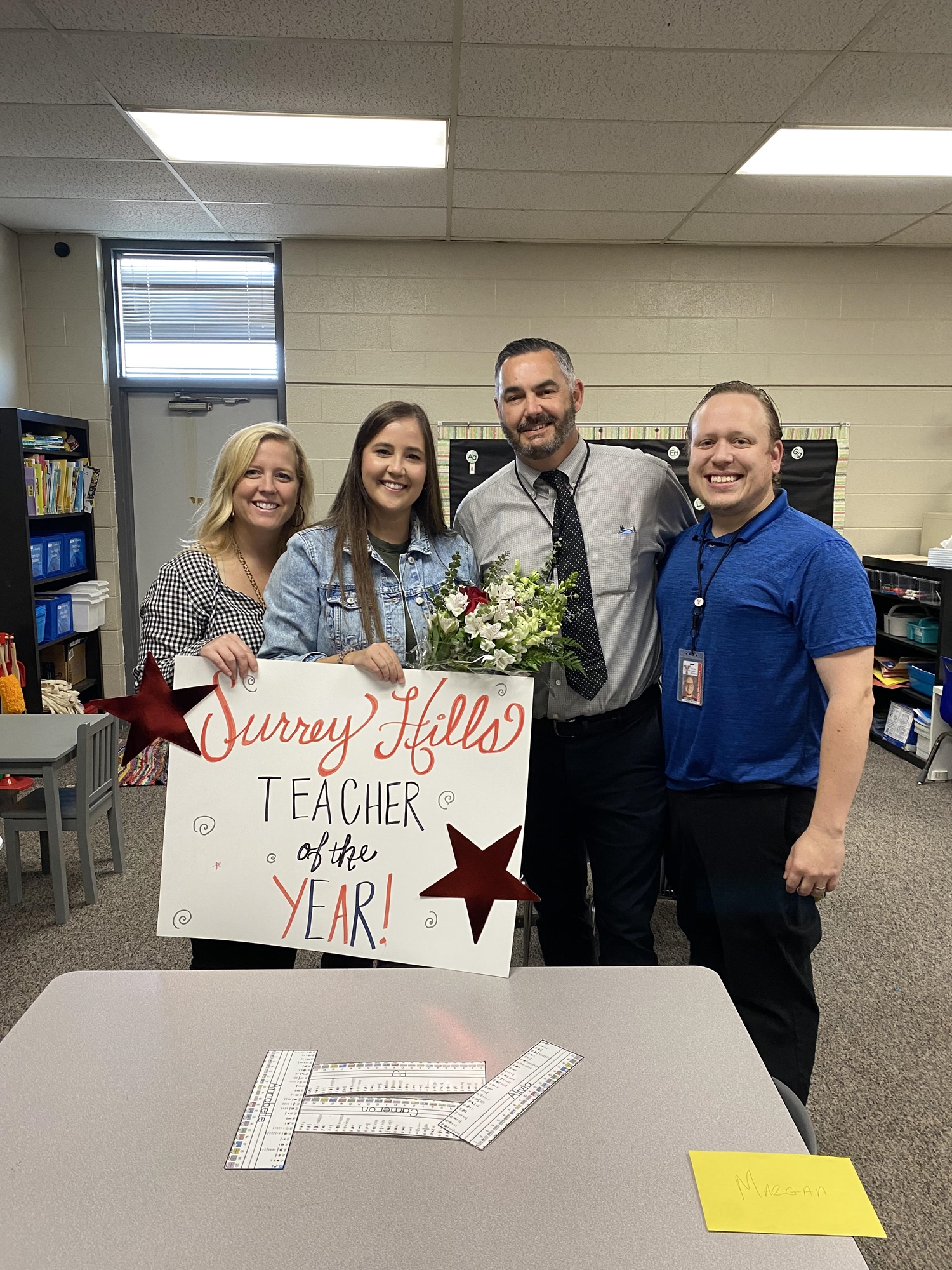 A group of four people, including a teacher holding flowers and a sign that reads 'Sunny Hills Teacher of the Year,' pose together in a classroom setting.