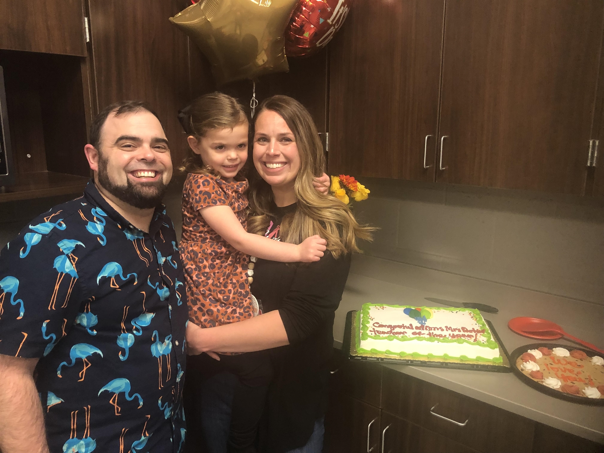 A smiling family of three poses together in a kitchen, with a birthday cake and cookies on the counter, and balloons in the background.