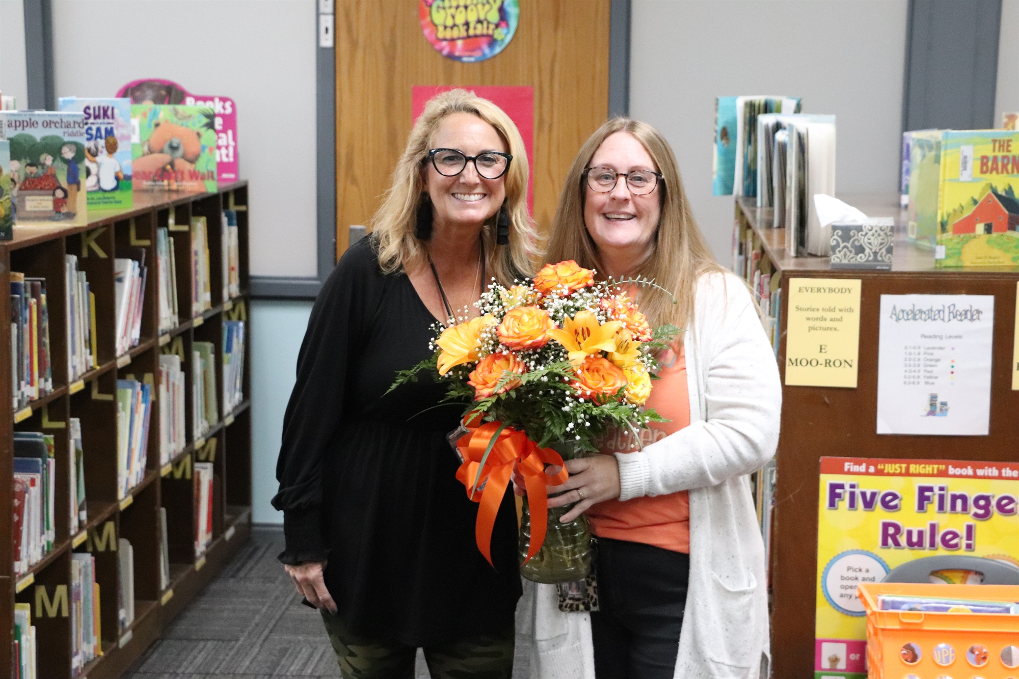 Two women stand together in a library, one holding a bouquet of orange flowers, with shelves of books in the background.