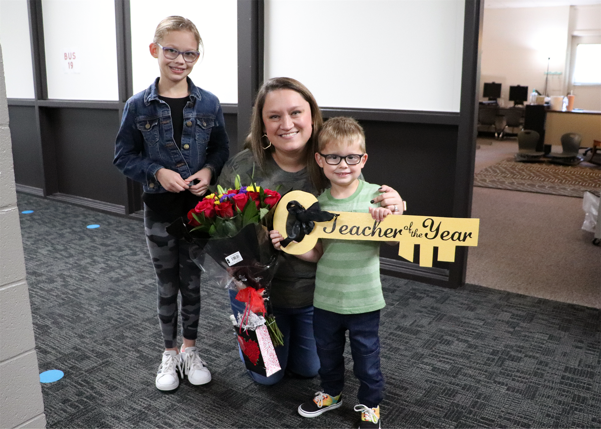 A smiling teacher holds a large key labeled 'Teacher of the Year' while standing between two children, one holding a bouquet of flowers, in a school hallway.