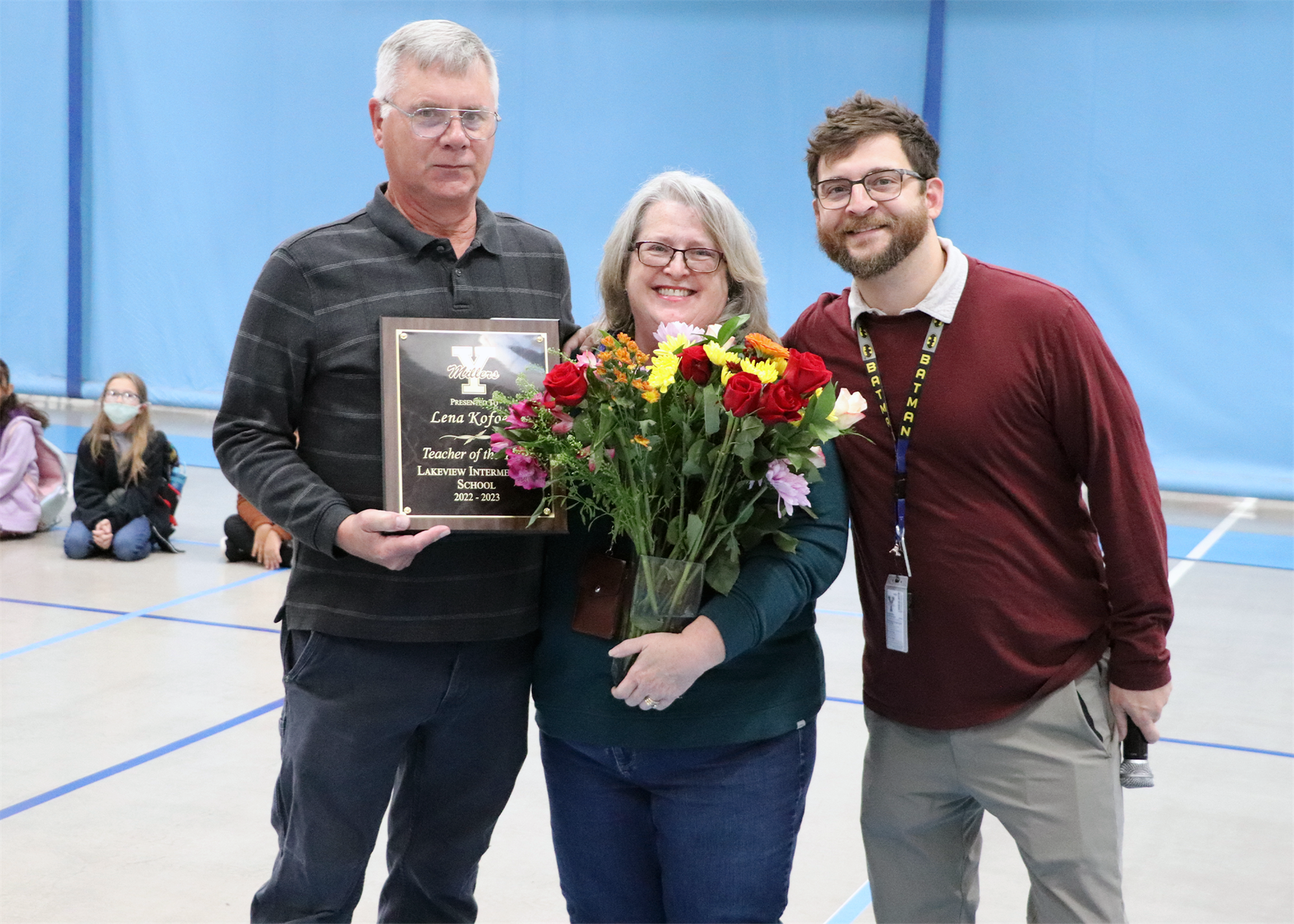Three individuals stand together in a gymnasium, with one man holding a plaque, a woman in the center holding a bouquet of flowers, and another man smiling beside them.