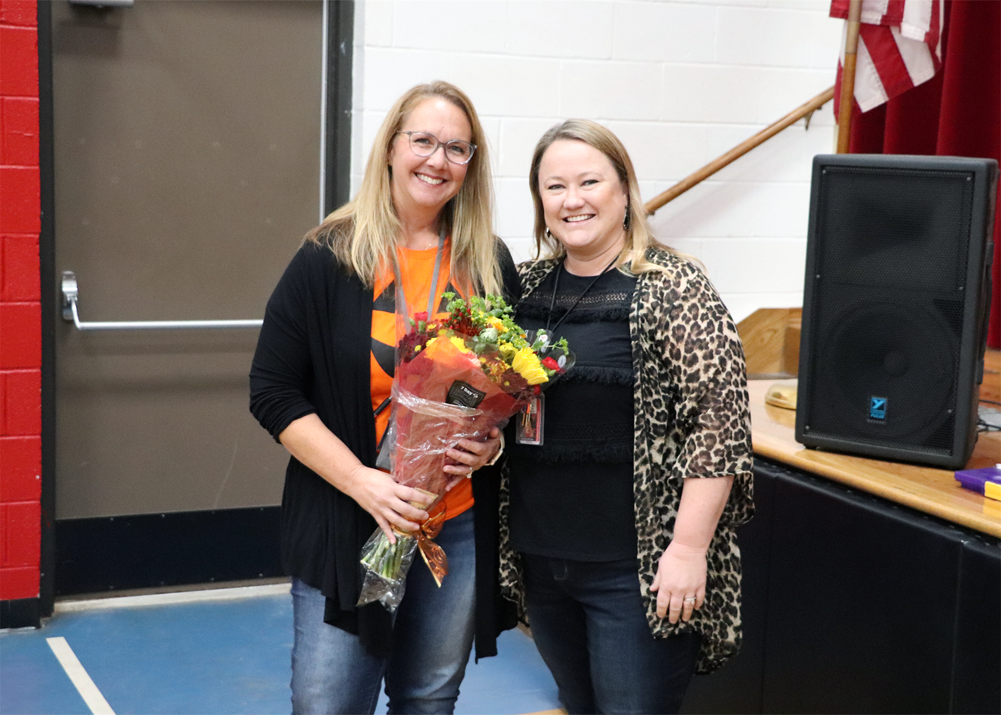 Two women stand together, one holding a bouquet of flowers, in a school setting with a microphone and American flag in the background.