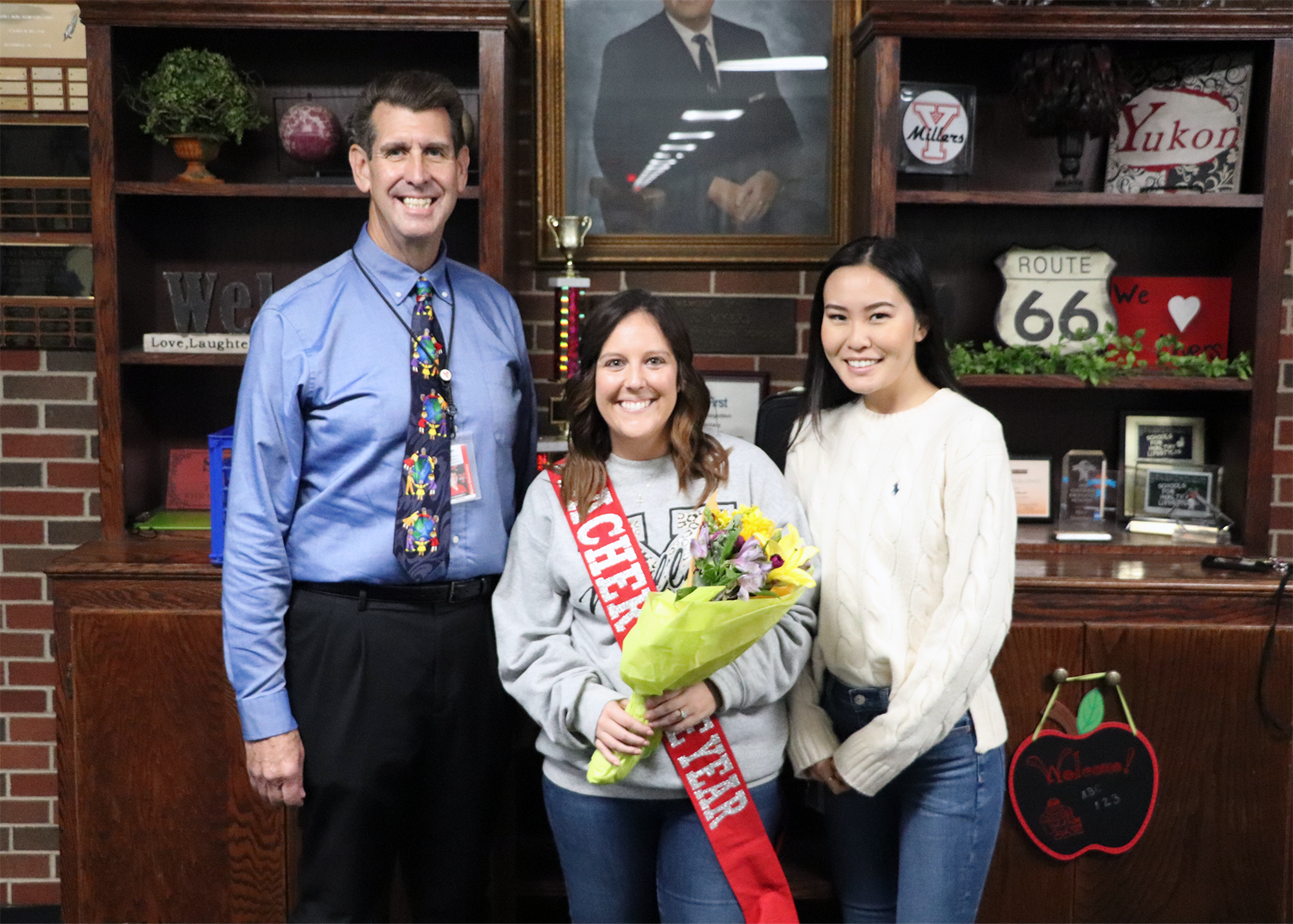 A group of three people, including a woman holding a bouquet and wearing a sash, pose for a photo in front of a decorated shelf with a portrait and various items.
