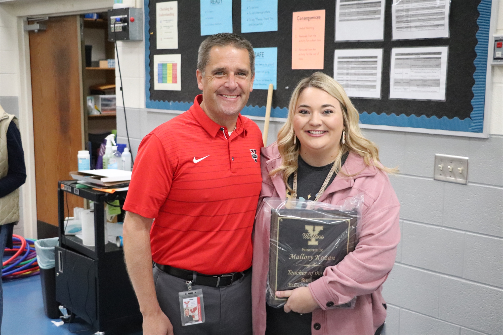A man in a red shirt and a woman in a pink sweater stand together, smiling while holding an award plaque in a classroom setting with educational materials on the walls behind them.