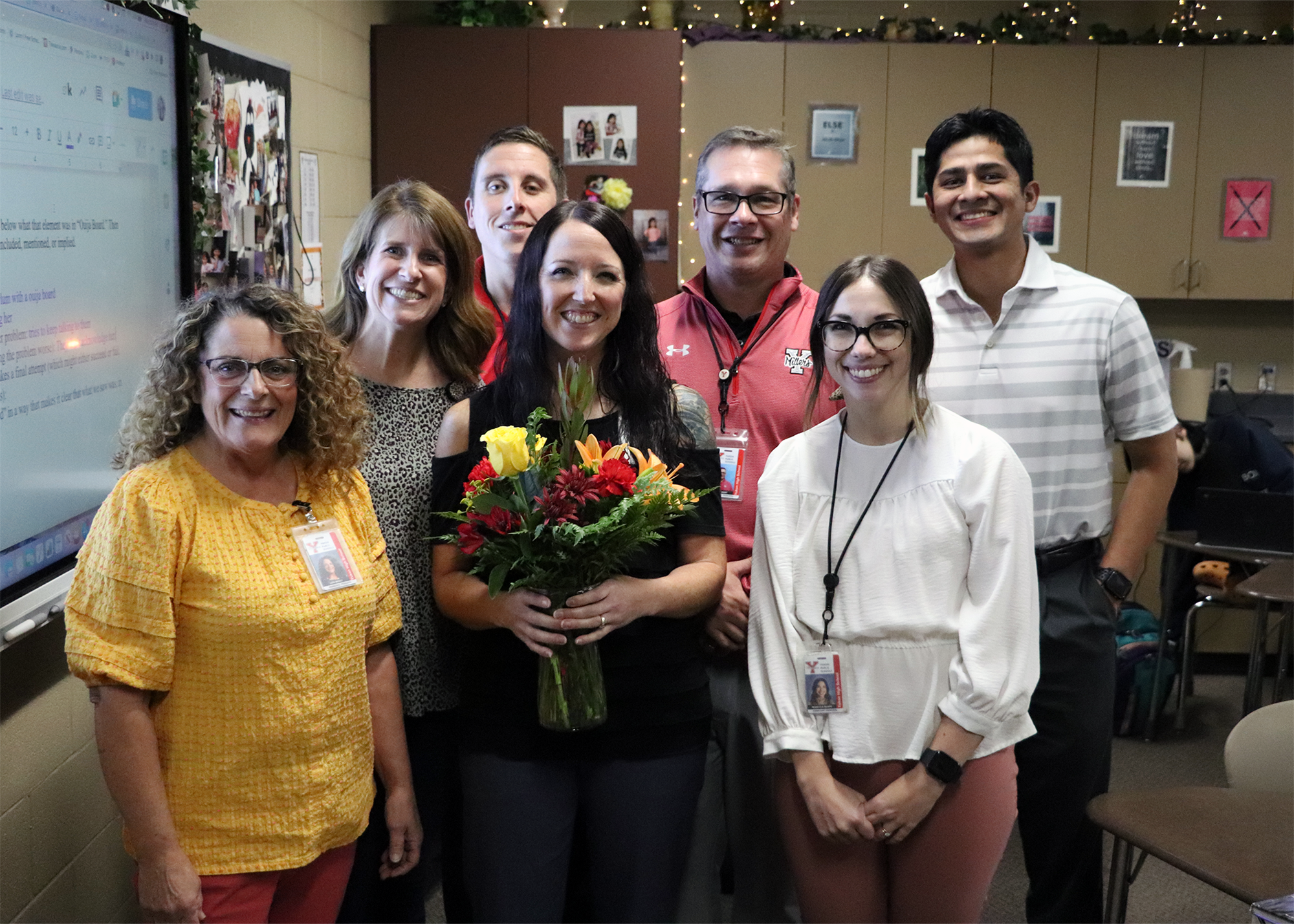 A group of seven people, including a woman holding a bouquet of flowers, pose together in a classroom setting, smiling at the camera.