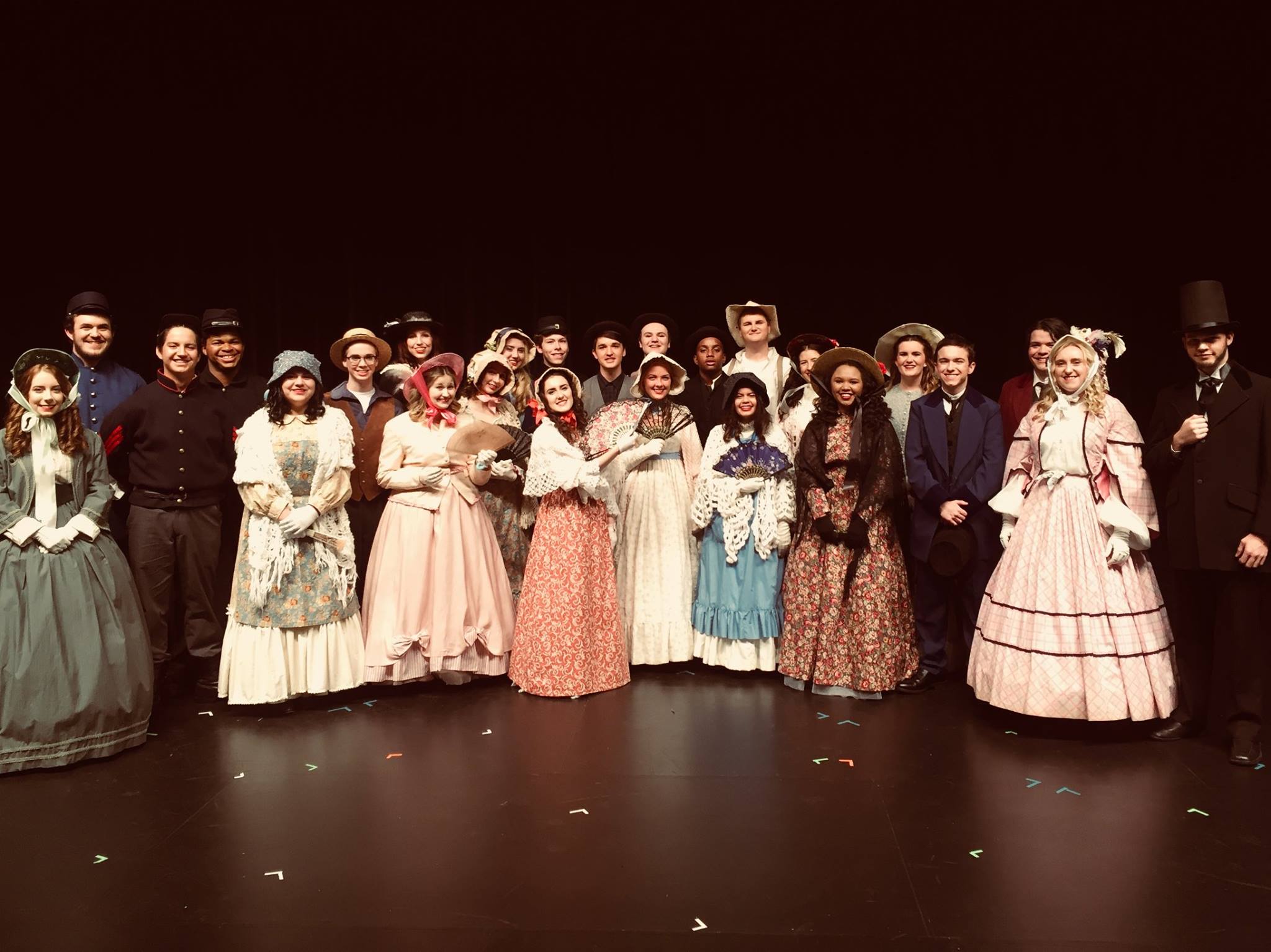 A large group of performers dressed in historical costumes poses together on stage, showcasing a variety of outfits from different eras, with a dark background.
