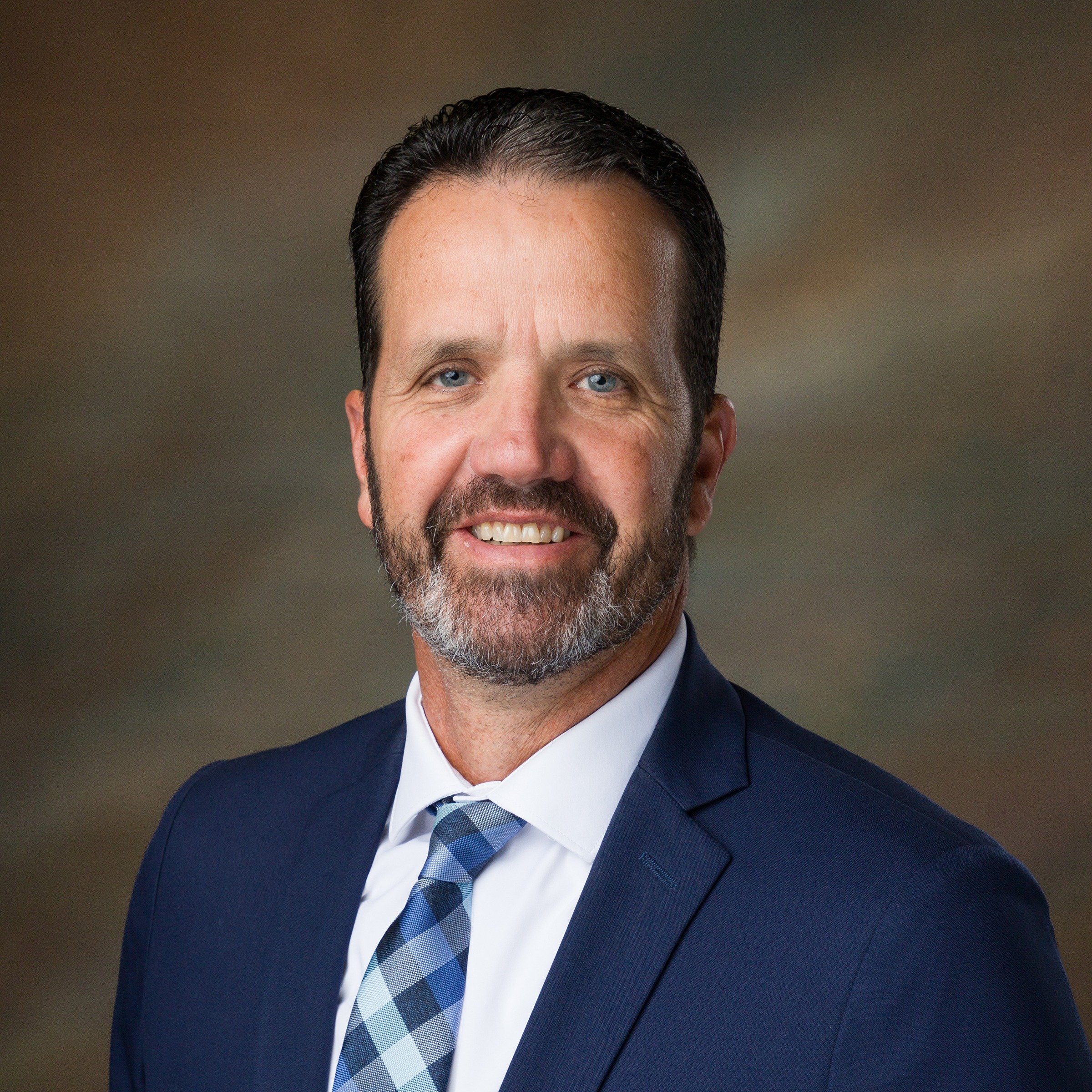 A smiling man with facial hair and short gray hair, wearing a white shirt and a  plaid tie.