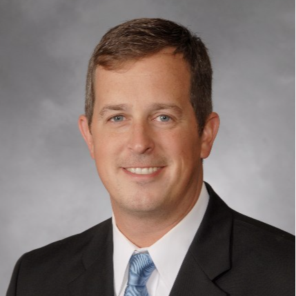 A professional headshot of a man in a black suit and a light blue tie, smiling against a gray background.