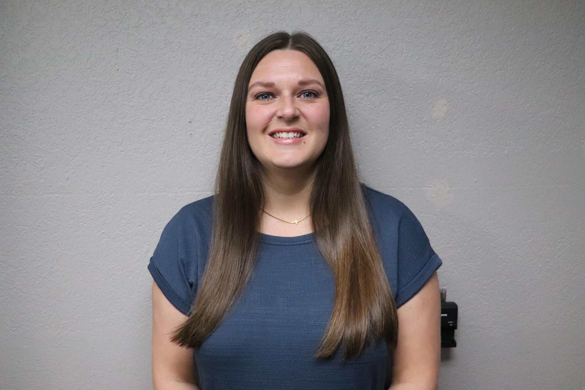 a girl with brown hair smiling in front of a gray wall with a blue shirt