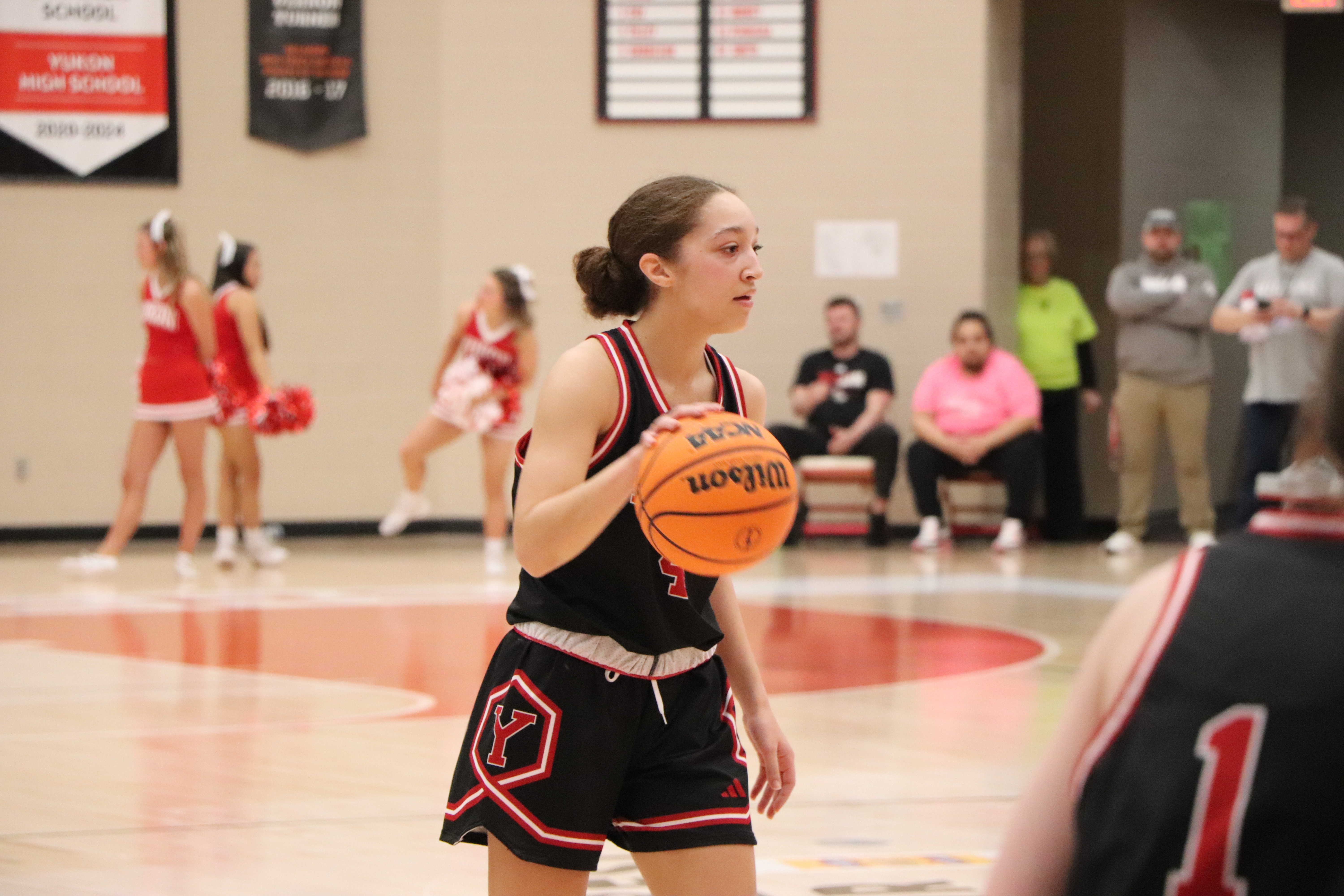 A female basketball player in black and red uniform dribbles a Wilson basketball on a gym court. Cheerleaders and spectators in the background.