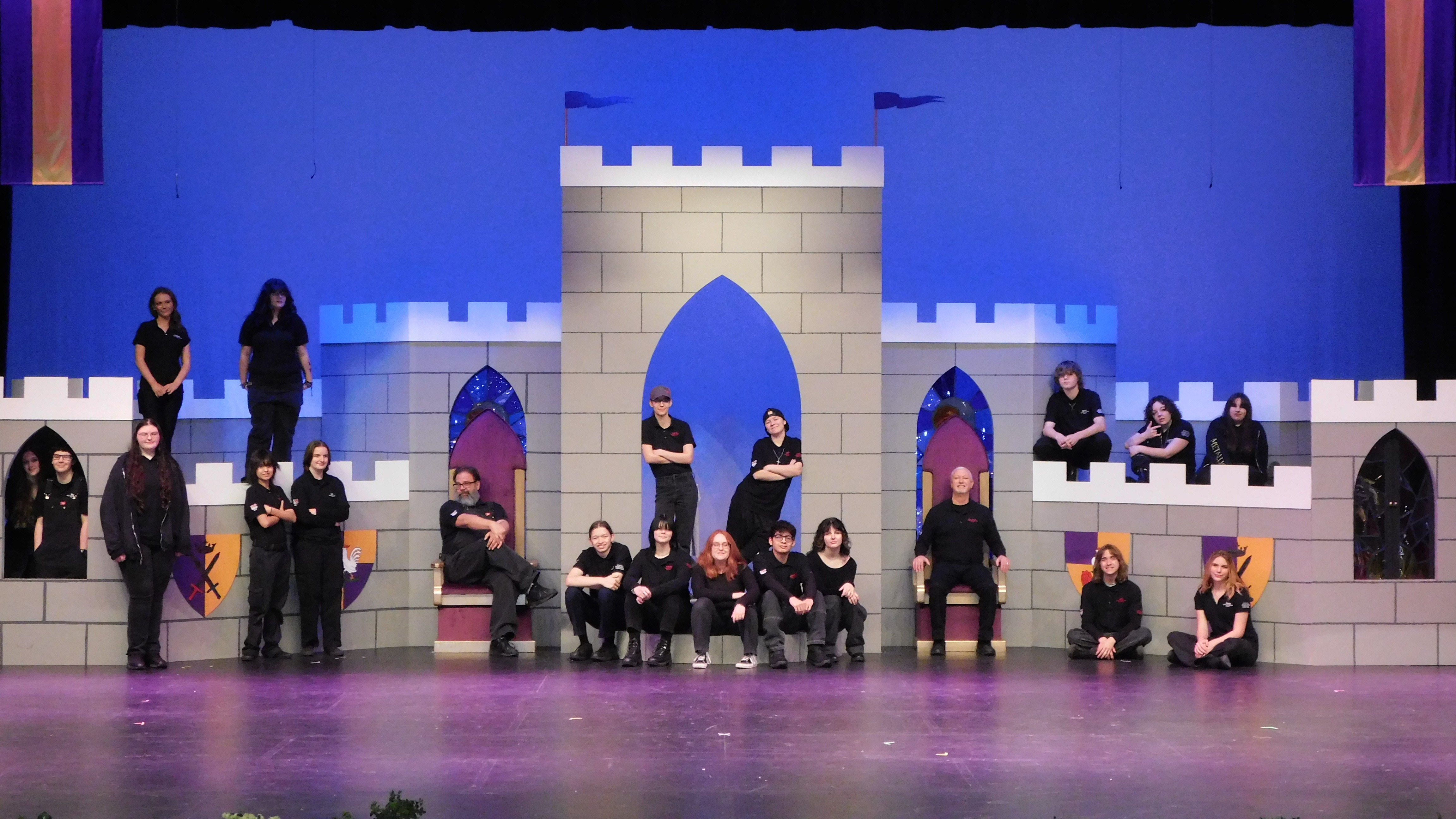 A group of students and instructors pose together on stage, dressed in black shirts, with a colorful backdrop of a medieval castle behind them.