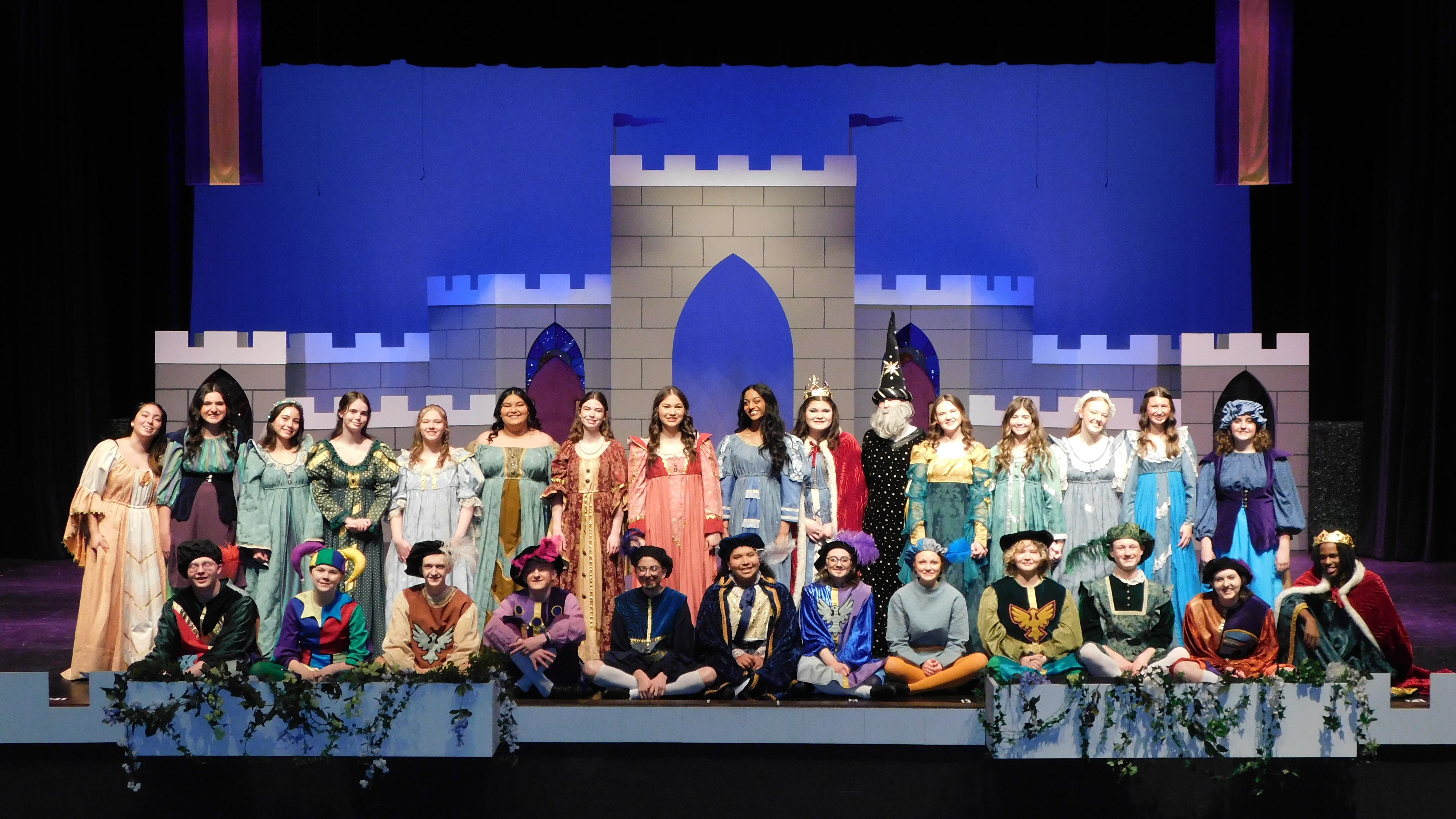 A large group of performers dressed in historical costumes poses together on stage, showcasing a variety of outfits from different eras, with a dark background.