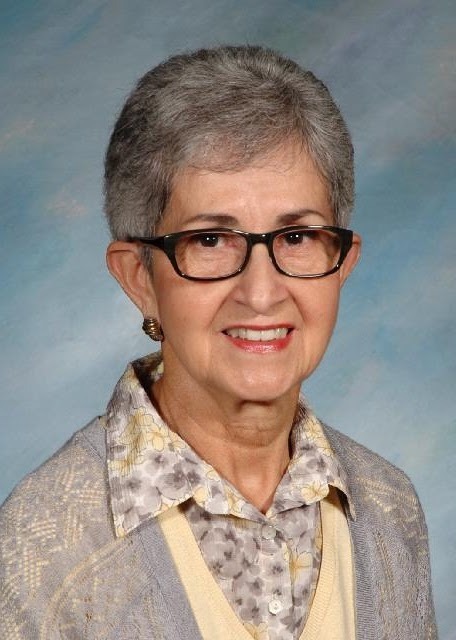 Woman with dark grey hair wearing glasses and a floral grey and yellow top along with a grey sweater