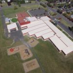This is an aerial view of a large, rectangular building with red roofs, possibly a school or community center, surrounded by open grassy areas.