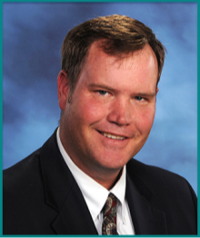 A professional headshot of a man in a suit and tie, smiling against a soft blue background.