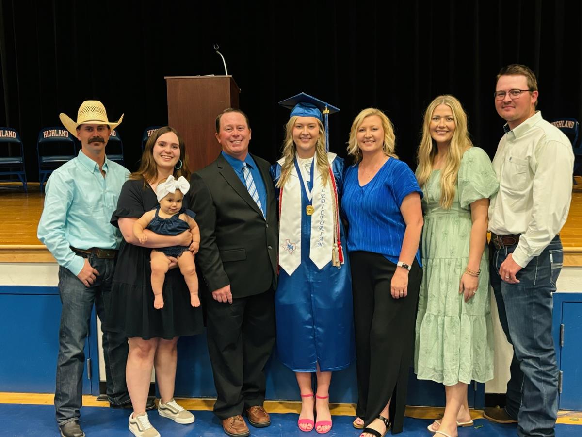 A group of seven people, including a graduate in a blue cap and gown, pose together for a photo at a graduation ceremony, with a podium in the background.