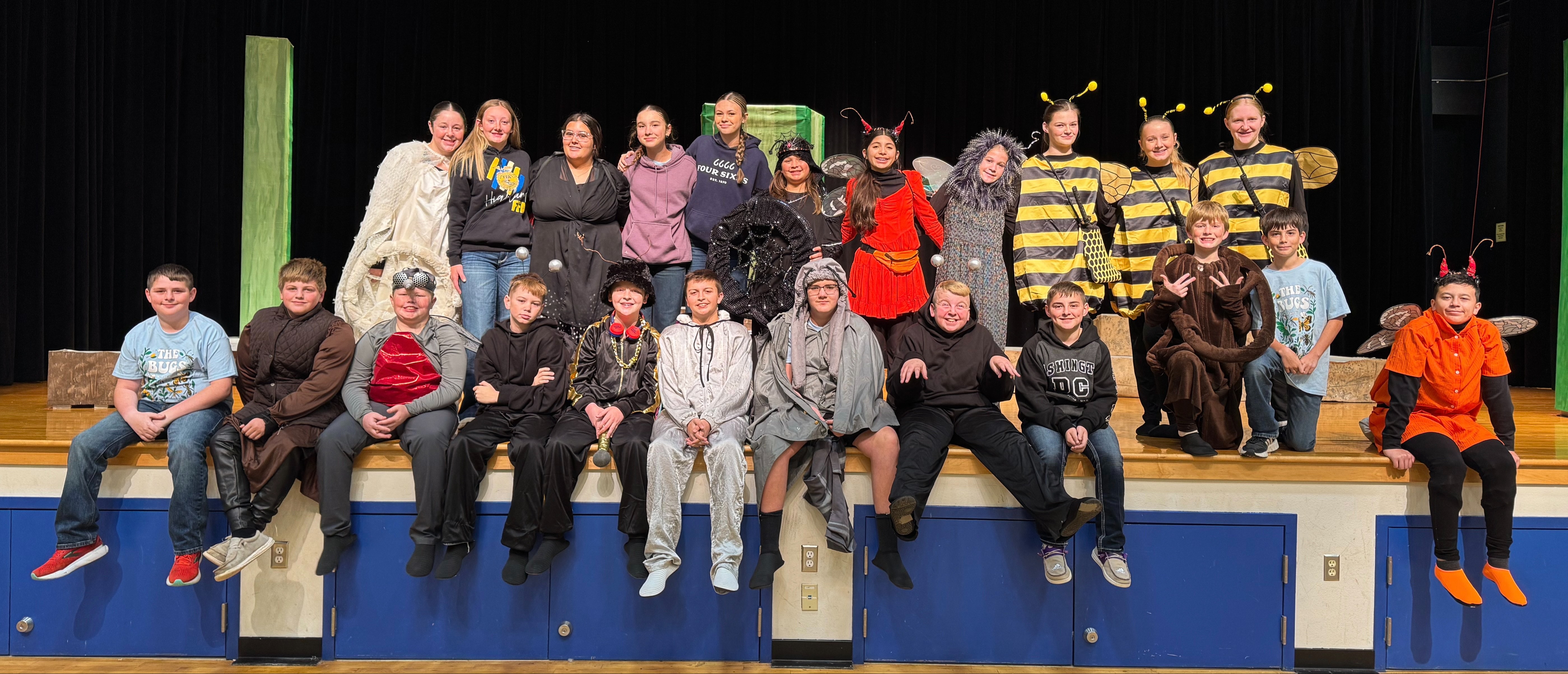 A group of children and adults dressed in various costumes, including animals and characters, posing together on a stage with a black curtain in the background.