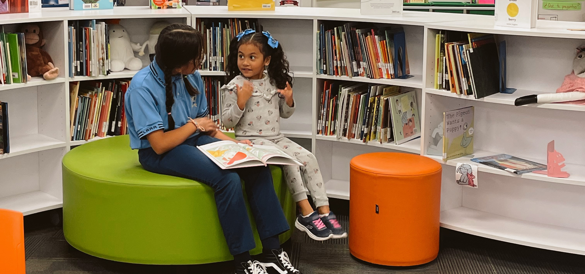 A young girl sitting on a stool, engaged in conversation with an adult in a library setting.