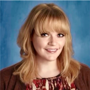 A woman with blonde hair and bangs smiles in front of a blue background, wearing a patterned top and a cardigan.