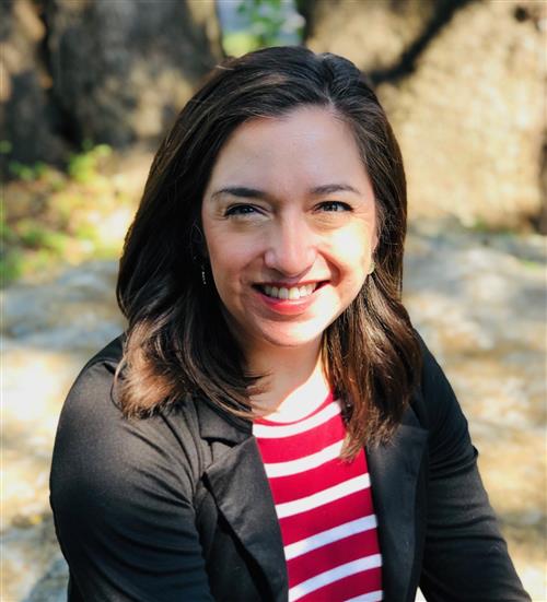 A woman with shoulder-length brown hair smiles at the camera while sitting outdoors, wearing a black jacket over a red and white striped shirt, with trees and rocks in the background.