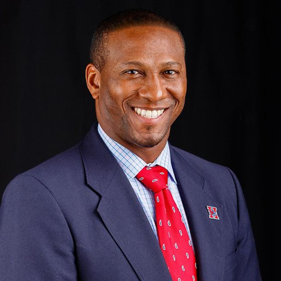 A smiling man in a dark suit and red tie stands against a black background, exuding a professional demeanor.
