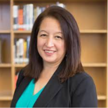 A woman with shoulder length brown hair, wearing  a black business jacket over a light blue shirt while smiling at the camera.with bookshelves in the background.