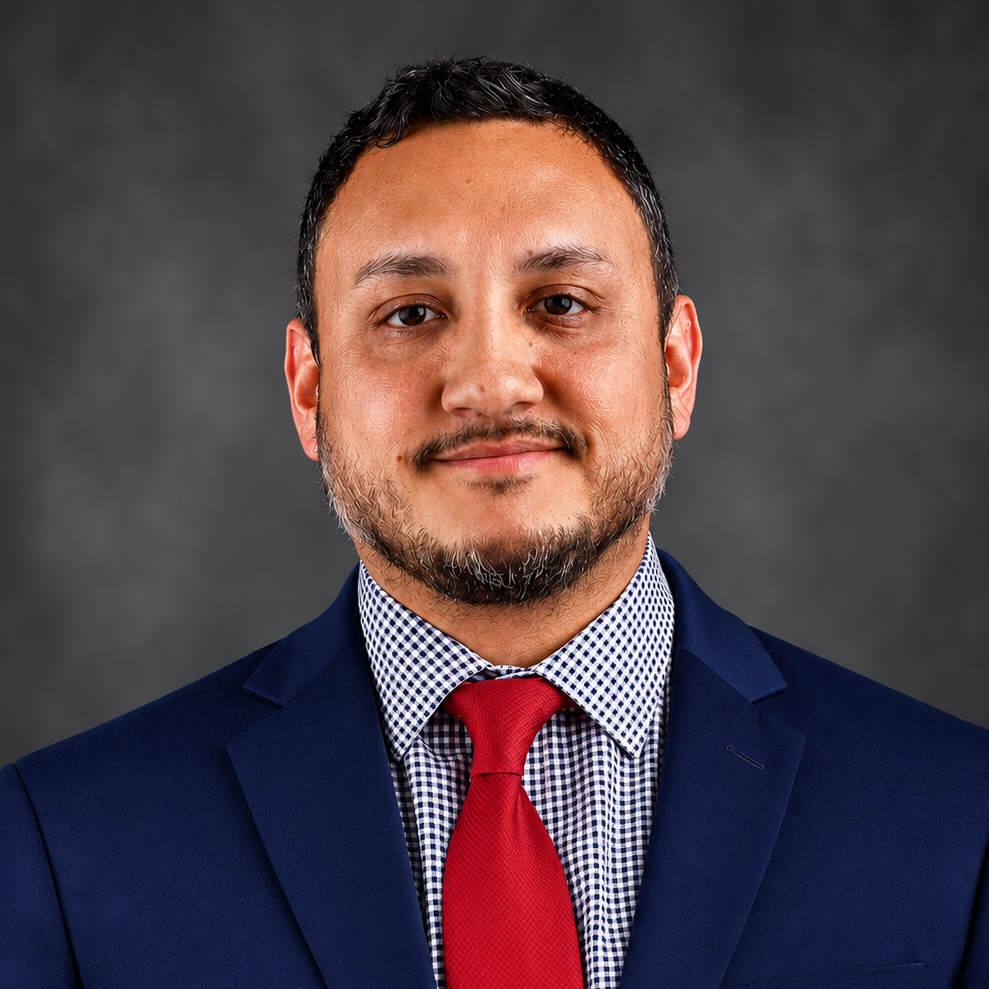 A man with short black hair, wearing a dress shirt and red tie beneath a navy blue business jacket while standing in front of a dark gray professional background.