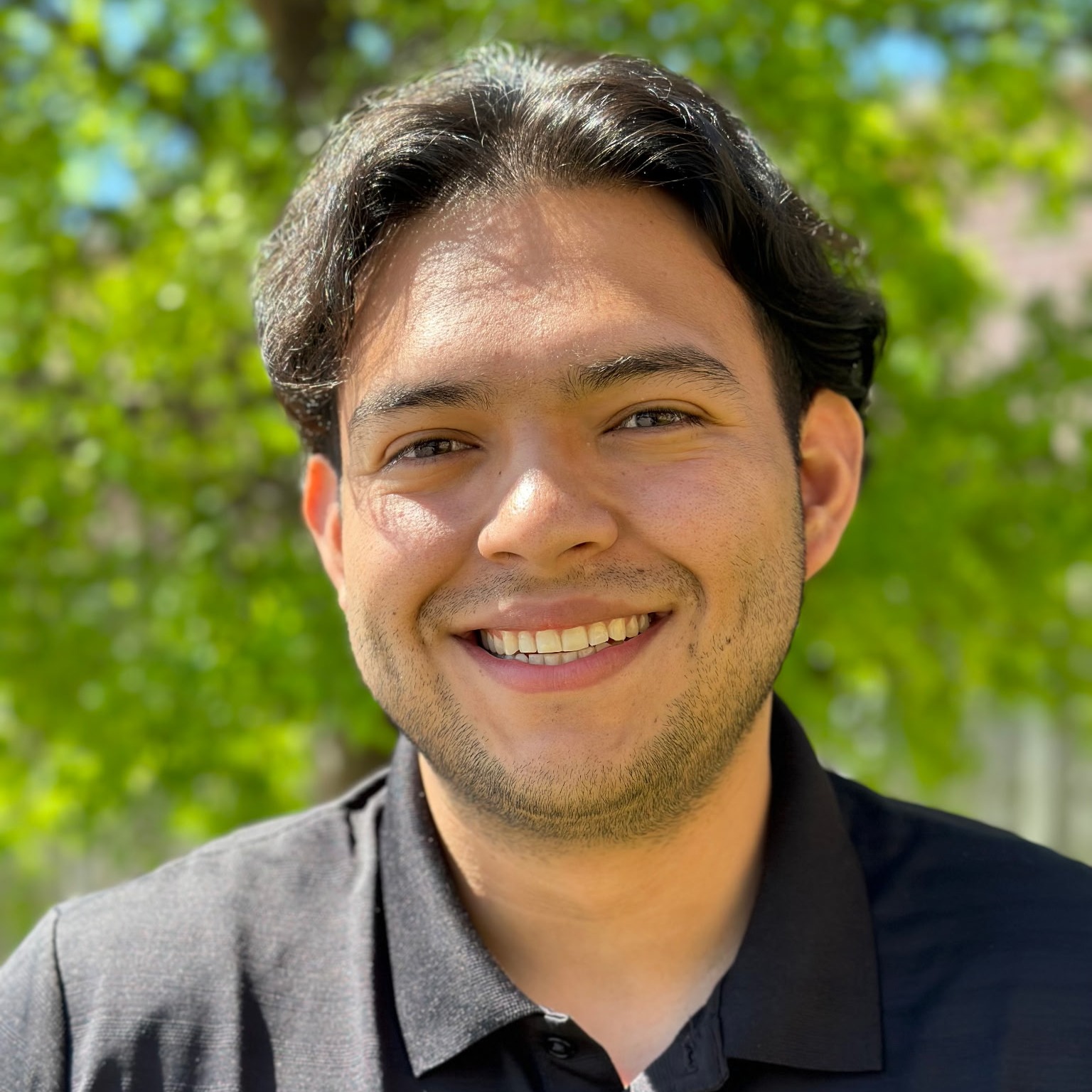 A man in a black collared shirt smiling with trees in the background