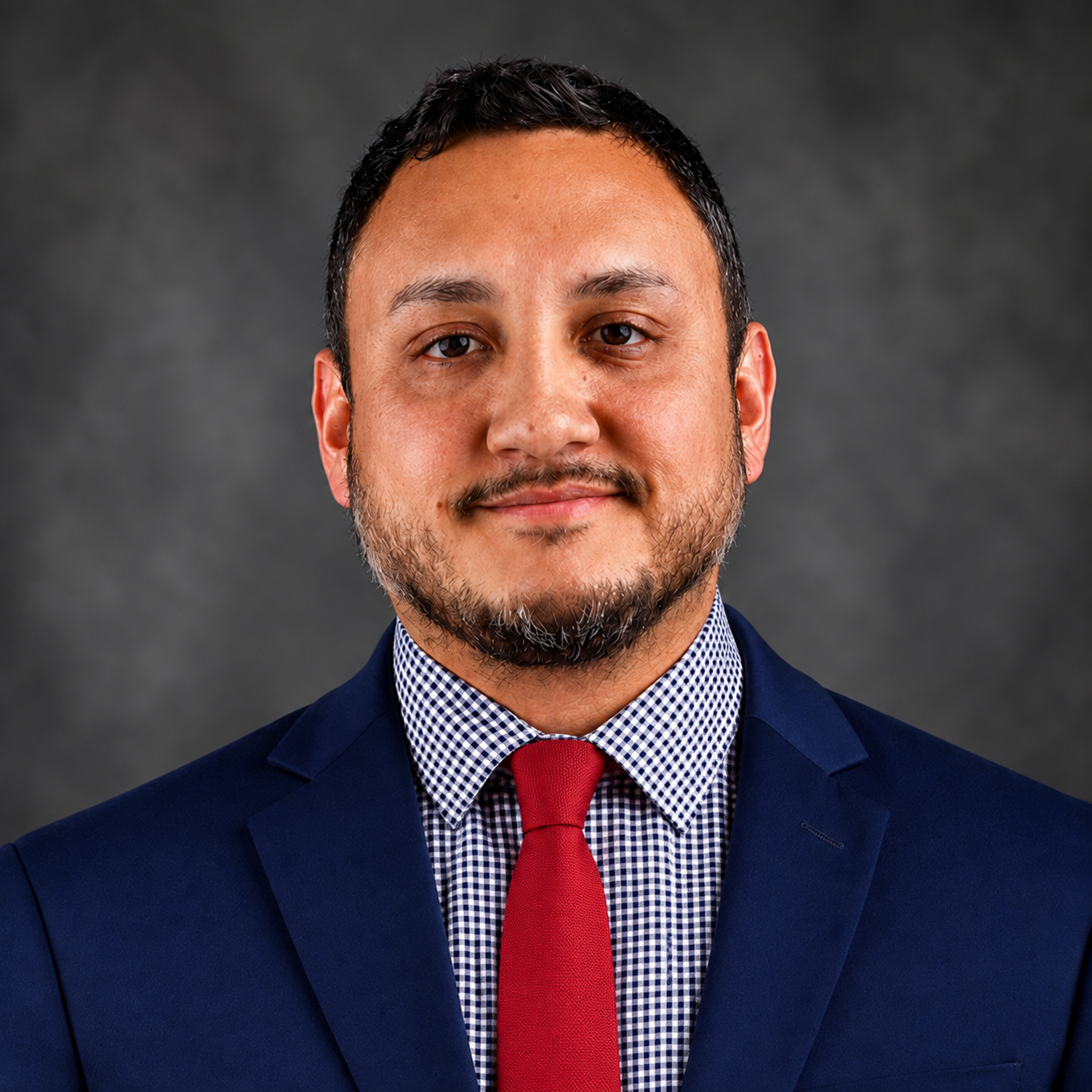 A man with short black hair, wearing a dress shirt and red tie beneath a navy blue business jacket while standing in front of a dark gray professional background.