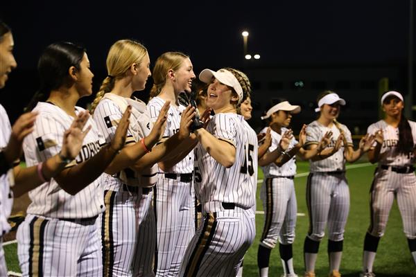 A group of female softball players in striped uniforms celebrate and cheer for a teammate on a field during the evening.