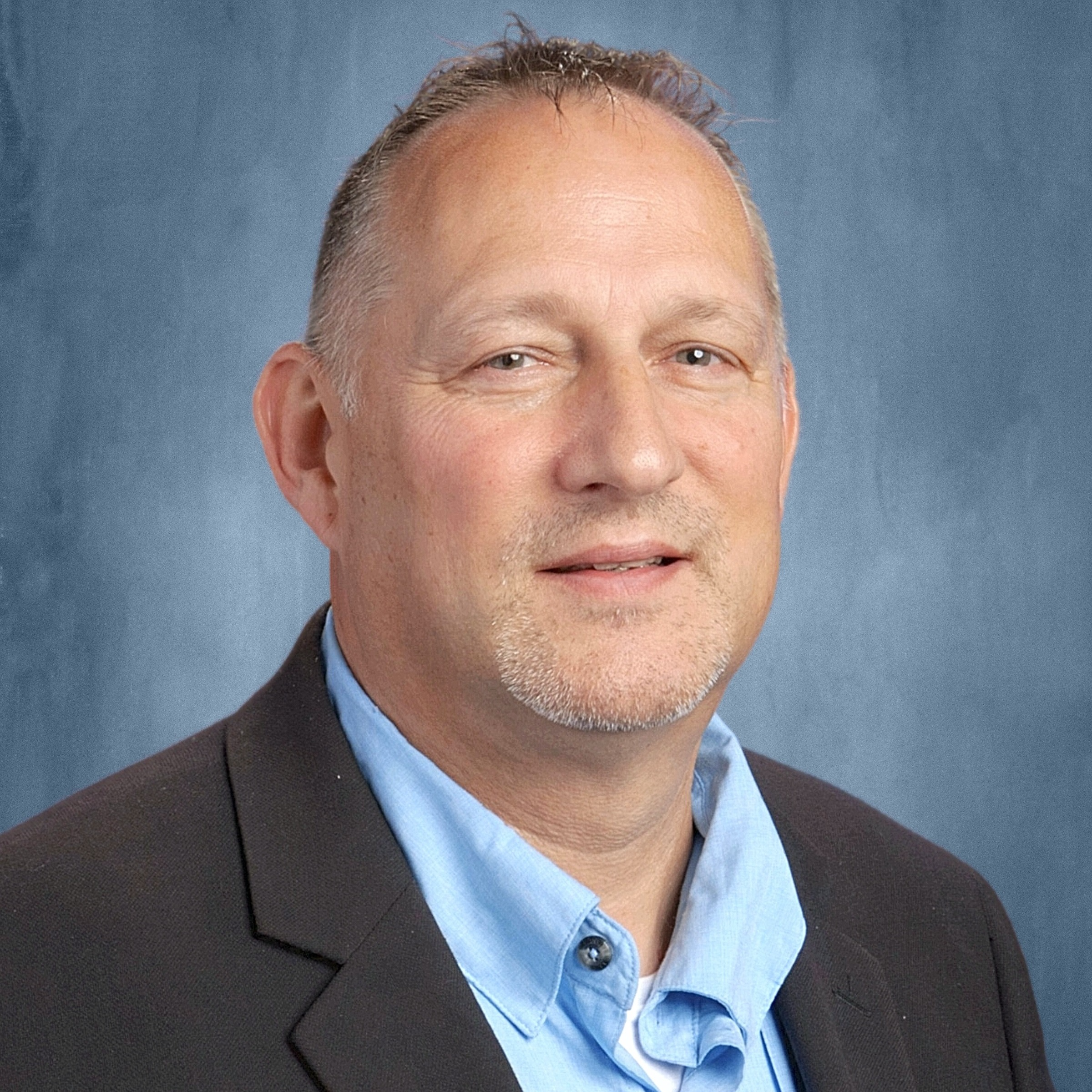 A man with short hair smiling at the camera, wearing a dark grey jacket with a blue shirt underneath while against a dark blue photo background.