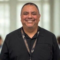 A man with very short hair smiling at the camera, while wearing a black button up shirt with a lanyard around his neck.