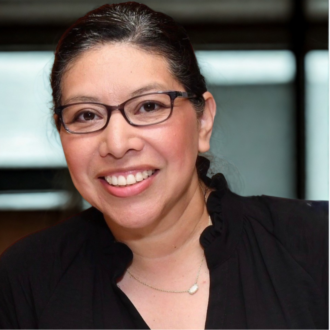 A woman with pulled back hair in glasses and a black shirt smiling at the camera against an out of focus background.