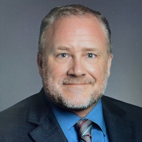 A man with short hair and beard wearing a dark grey jacket with a blue shirt and grey tie with maroon and blue accents, grinning at the camera against a grey background.