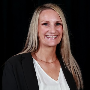 A woman with long blonde hair in a black jacket with a white shirt underneath smiling at the camera against a black background.