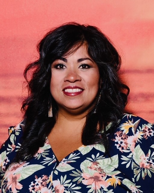 A woman with dark, wavy hair wearing a floral-patterned shirt smiles against a sunset background.