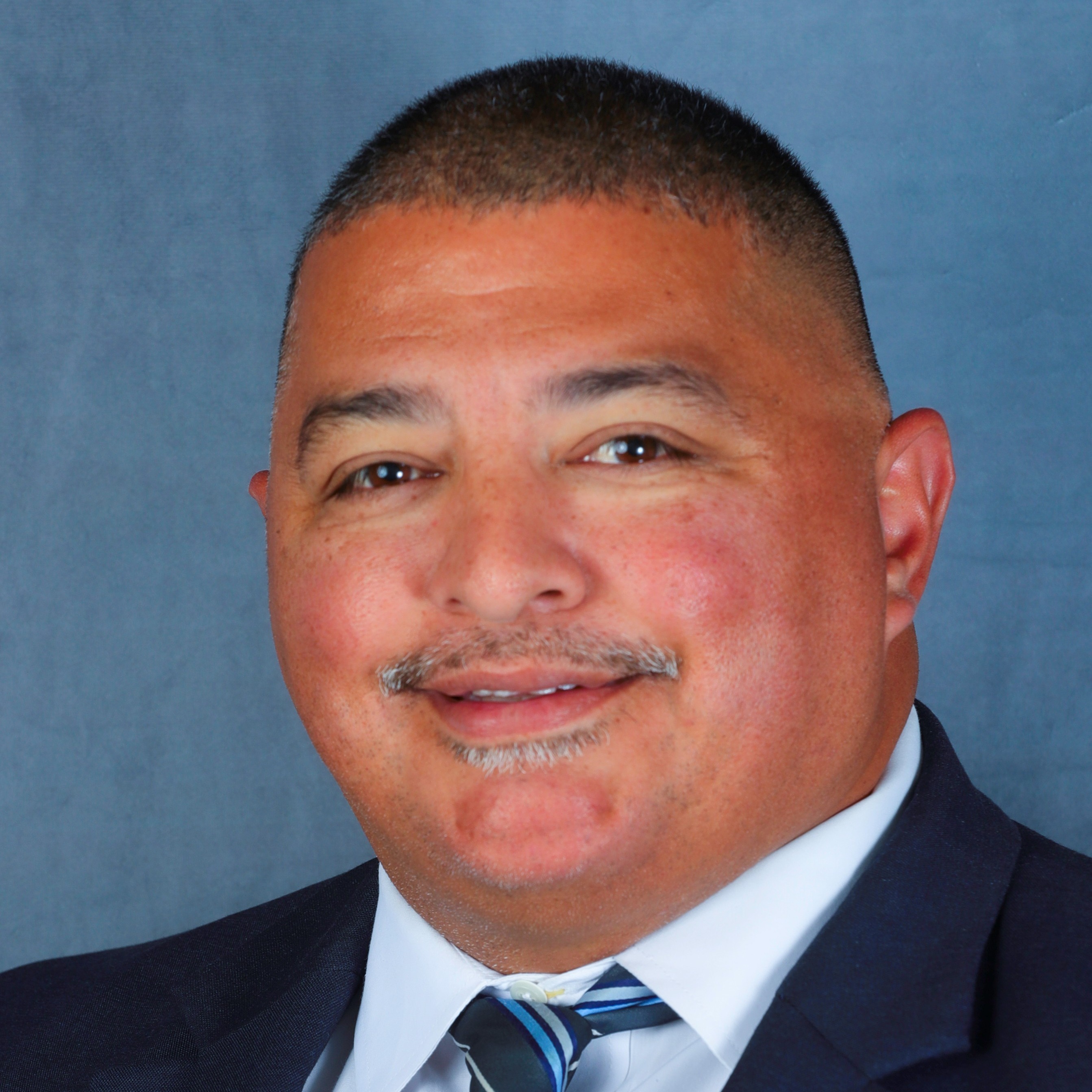A man with short hair and mustache wearing a jacket with a white shirt and blue tie smiling at the camera against a blue background.