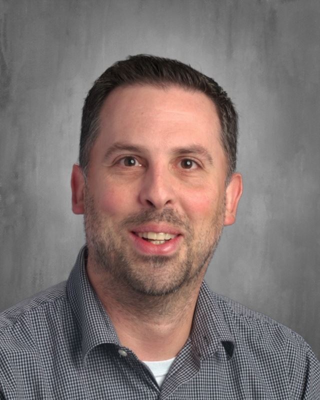 A smiling man with short dark hair and a beard, wearing a checkered shirt, poses for a portrait against a gray background.