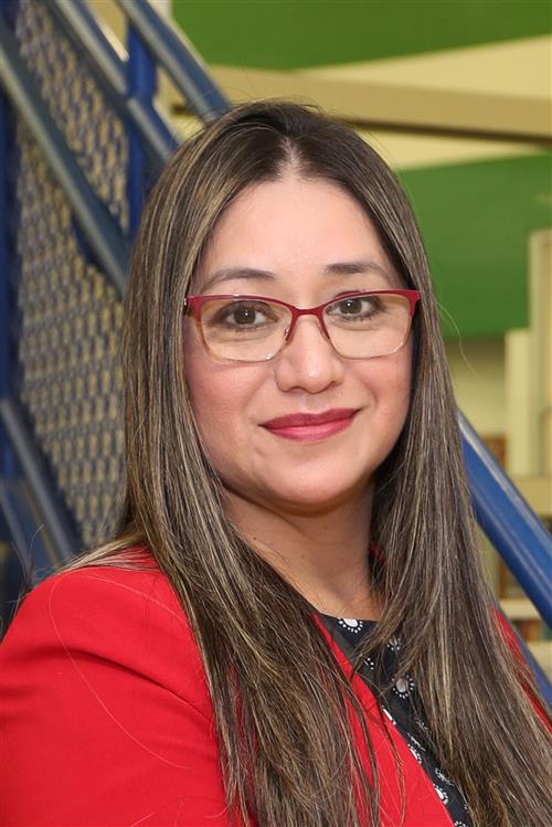 A woman with long, straight hair wearing glasses and a red blazer smiles in front of a green and blue background.