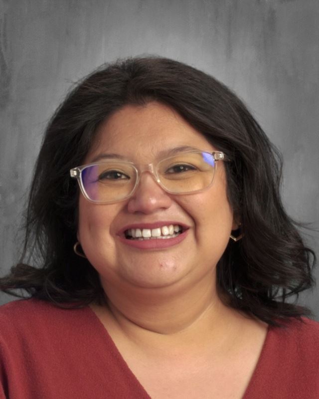 A smiling woman with long, dark hair and glasses, wearing a red top, against a neutral background.