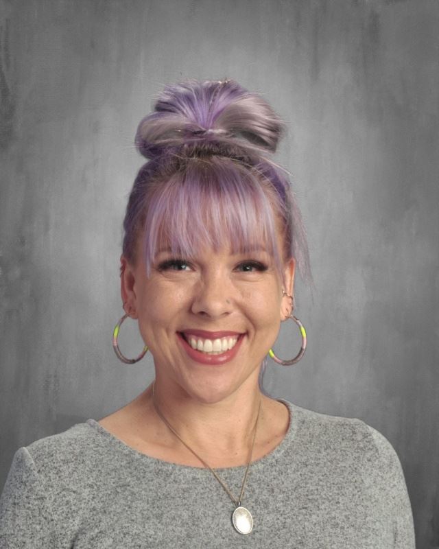A smiling woman with purple hair styled in a bun, wearing hoop earrings and a gray sweater, set against a textured gray background.